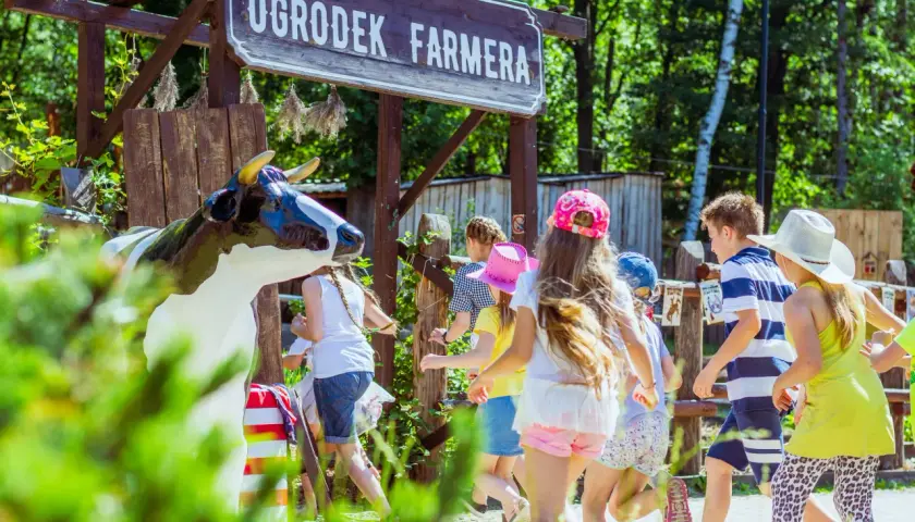 Dzieci biegną obok drewnianego szyldu "Ogródek Farmera" i figury krowy.